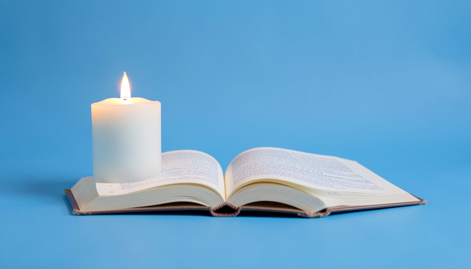 A single glowing candle beside an open book on a soft blue studio backdrop, calm and reflective mood